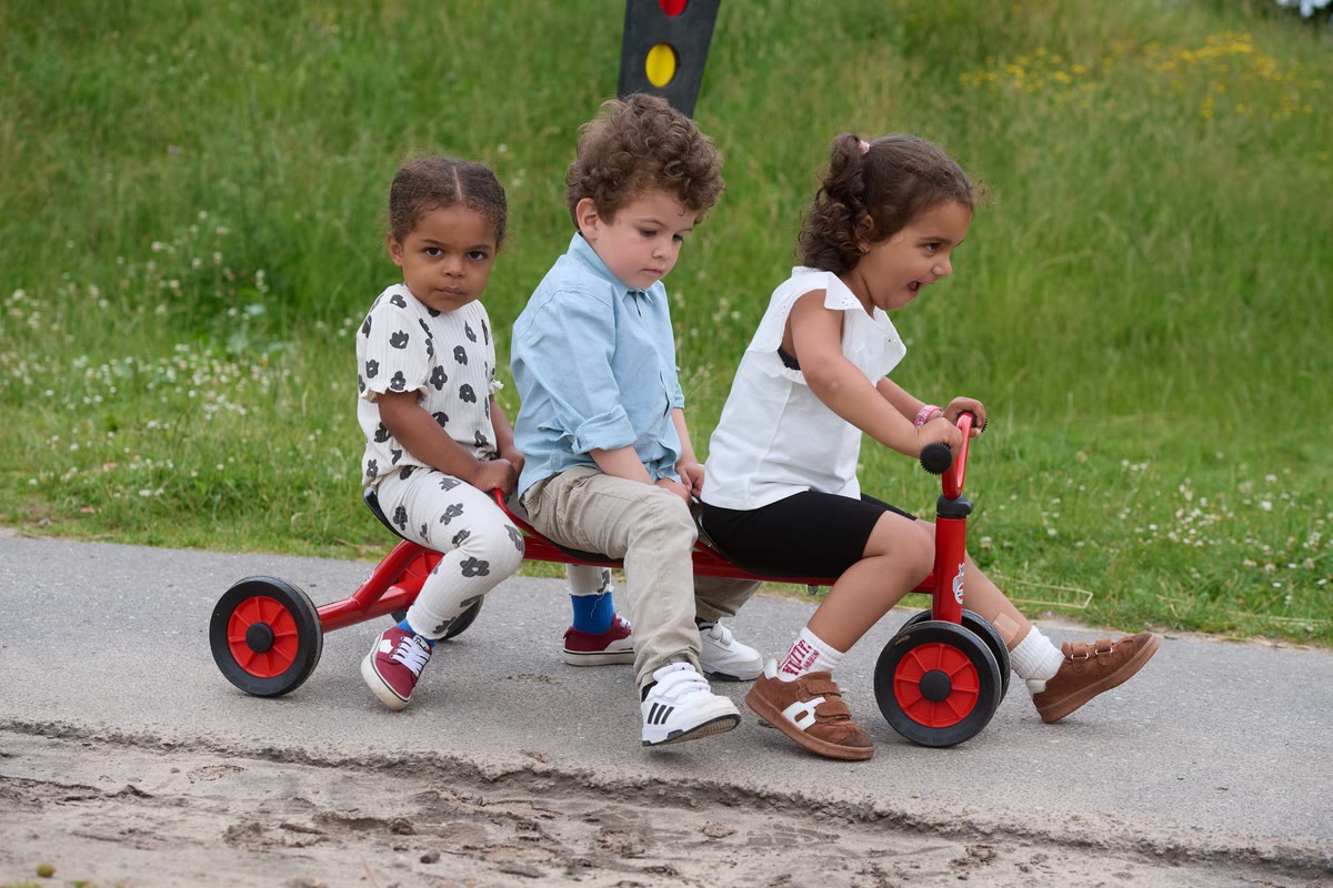Drei Kinder fahren gemeinsam auf einem roten Winther-Dreirad mit langer Sitzbank über einen Weg im Außengelände