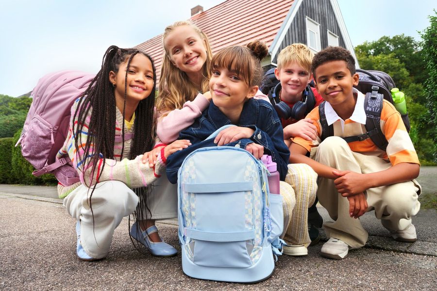 Fünf Kinder mit Satch-Schulrucksäcken hocken auf einem Gehweg vor einem Haus