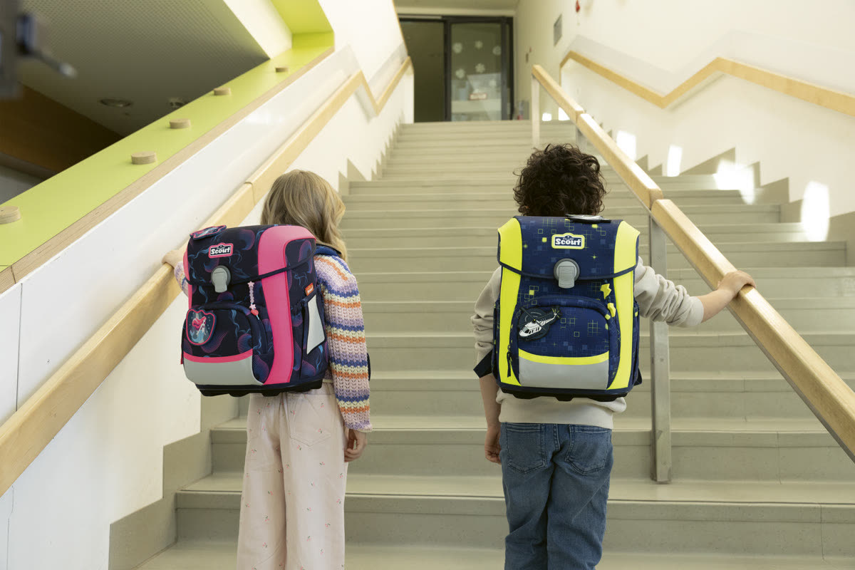 Zwei Kinder mit Scout Schulranzen gehen eine Treppe im Schulhaus hinauf, von hinten gesehen