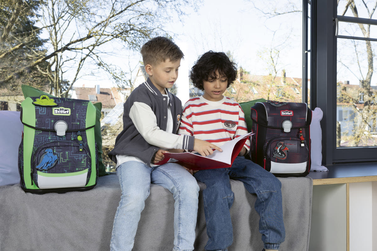 Zwei Jungen sitzen auf einer Bank und lesen gemeinsam in einem Heft, neben ihnen zwei Scout Schulranzen
