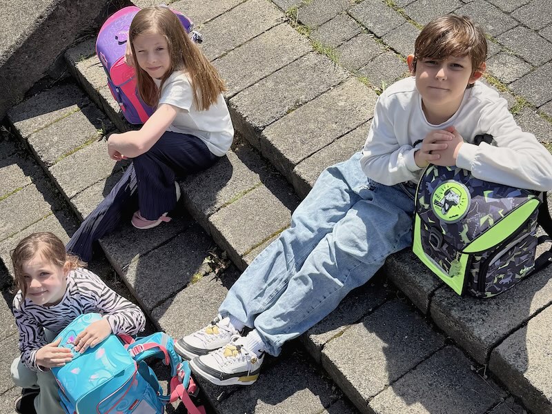 Drei Kinder sitzen mit ihren Schulranzen auf einer Treppe und schauen in die Sonne