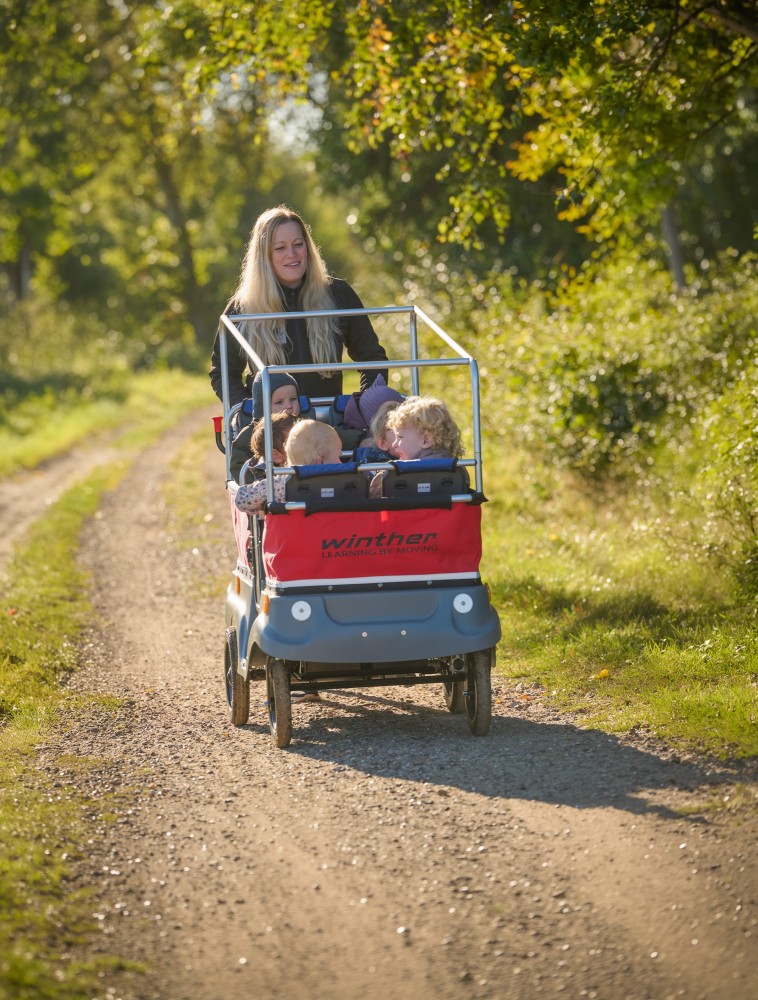 Erzieherin schiebt den Winther E-Turtle Kinderbus mit Motor für 6 Kinder mit Kindern einen Feldweg entlang
