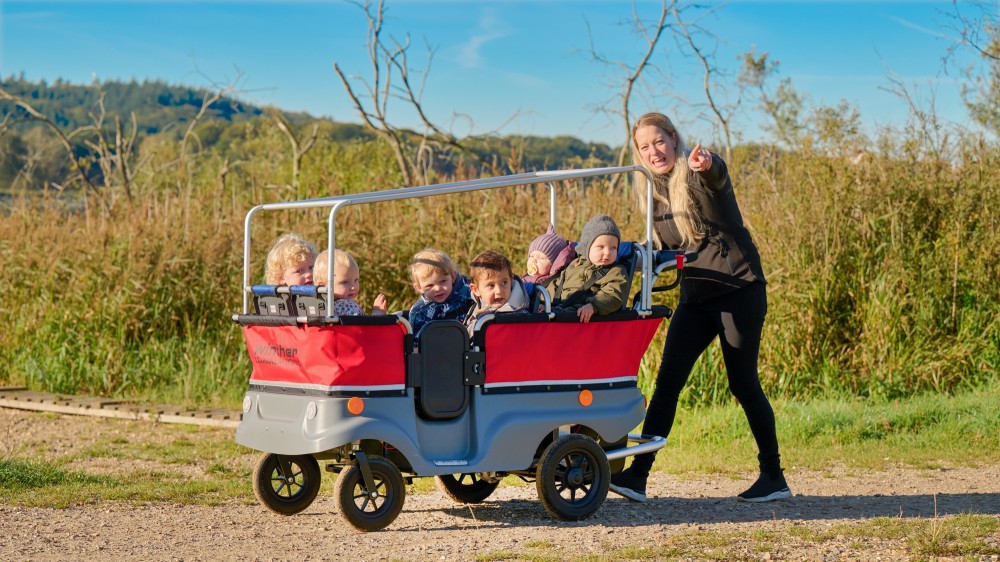 Erzieherin schiebt den Winther E-Turtle Kinderbus mit Motor für 6 Kinder mit Kindern durch die Natur